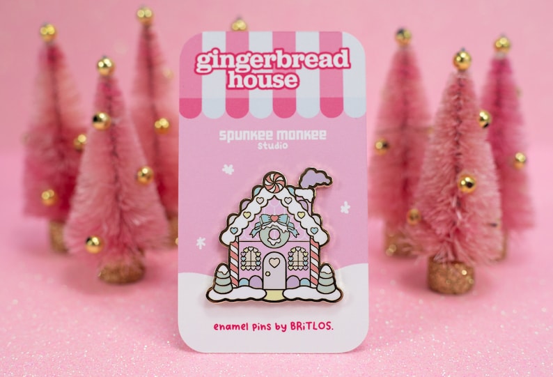 May include: A pink gingerbread house enamel pin with white frosting, candy canes, and a pink bow. The pin is on a pink card with the text "gingerbread house", "spunkee monkee studio", and "enamel pins by BRITLOS."