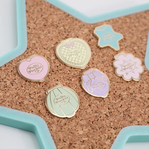 Puede incluir: Colecci&oacute;n de pines esmaltados con dise&ntilde;os de Polly Pocket en varias formas, incluyendo un coraz&oacute;n, estrella, flor y compactos. Los pines son de colores pastel con detalles dorados, colocados sobre una tabla de corcho dentro de un marco en forma de estrella azul claro.