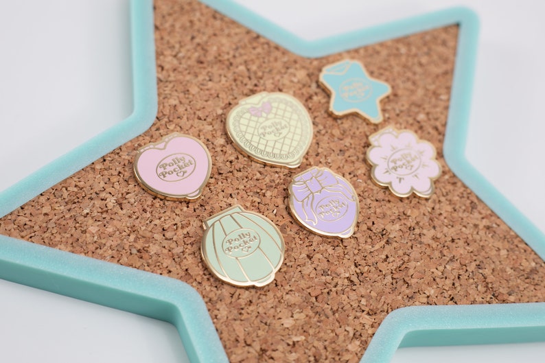 Puede incluir: Colecci&oacute;n de pines esmaltados con dise&ntilde;os de Polly Pocket en varias formas, incluyendo un coraz&oacute;n, estrella, flor y compactos. Los pines son de colores pastel con detalles dorados, colocados sobre una tabla de corcho dentro de un marco en forma de estrella azul claro.