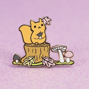 May include: A gold enamel pin featuring a cartoon squirrel holding a nut, sitting on a tree stump with mushrooms and leaves.