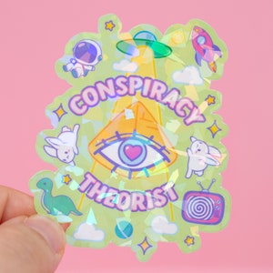 May include: A holographic sticker with a green background and a pyramid with an eye in the center. The text "CONSPIRACY THEORIST" is written around the pyramid. The sticker also features other images, including a rocket, a spaceship, a dinosaur, and a television.