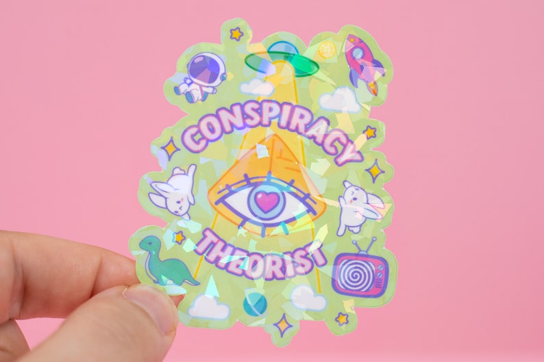 May include: A holographic sticker with a green background and a pyramid with an eye in the center. The text "CONSPIRACY THEORIST" is written around the pyramid. The sticker also features other images, including a rocket, a spaceship, a dinosaur, and a television.