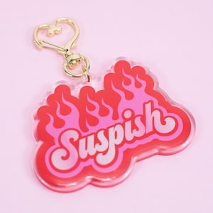 May include: A pink and red acrylic keychain with a gold heart-shaped clasp. The keychain features the word "Suspish" in a retro font with flames behind it.