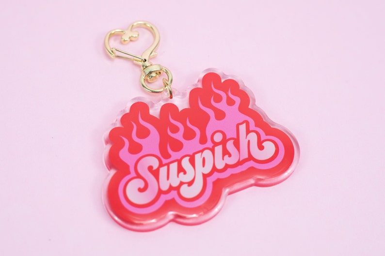 Pode incluir: Um chaveiro de acr&iacute;lico rosa e vermelho com um fecho dourado em forma de cora&ccedil;&atilde;o. O chaveiro apresenta a palavra "Suspish" em uma fonte retr&ocirc; com chamas atr&aacute;s.