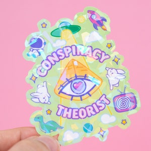 May include: A holographic sticker with a green background and a yellow pyramid with an eye in the center. The text "CONSPIRACY THEORIST" is written around the pyramid. Other images include a UFO, a rocket, a dinosaur, and bunnies.