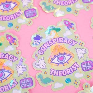 May include: A set of holographic stickers featuring a green pyramid with an all-seeing eye, a UFO, and the text "Conspiracy Theorist". The stickers also feature other designs, including a rocket, a dinosaur, a bunny, and a television.