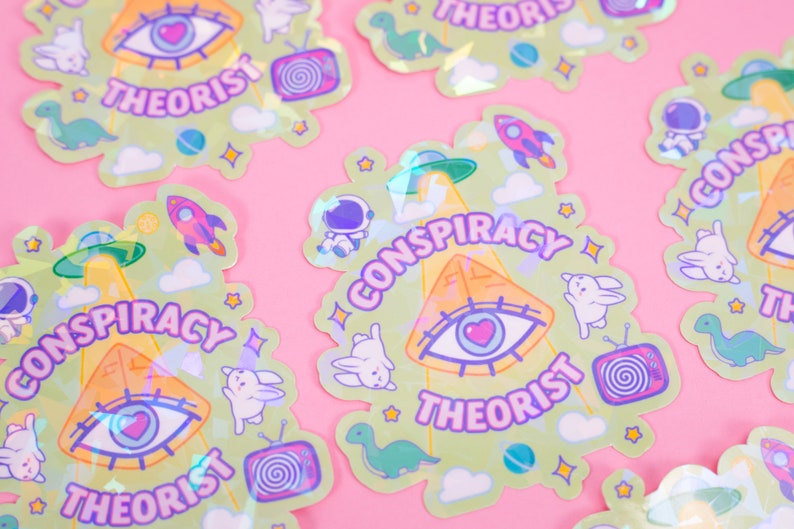 May include: A set of holographic stickers featuring a green pyramid with an all-seeing eye, a UFO, and the text "Conspiracy Theorist". The stickers also feature other designs, including a rocket, a dinosaur, a bunny, and a television.