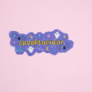 Puede incluir: Pegatina morada con un borde ondulado y un tema espeluznante. Presenta el texto "spooktacular" en letras amarillas, junto con fantasmas, murci&eacute;lagos y estrellas.