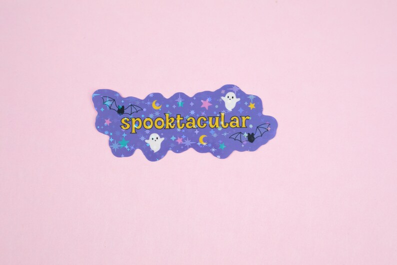 Puede incluir: Pegatina morada con un borde ondulado y un tema espeluznante. Presenta el texto "spooktacular" en letras amarillas, junto con fantasmas, murci&eacute;lagos y estrellas.