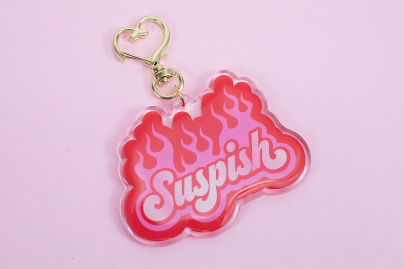 Pode incluir: Um chaveiro de acr&iacute;lico vermelho e rosa com um fecho dourado em forma de cora&ccedil;&atilde;o. O chaveiro apresenta a palavra "Suspish" em letras brancas com chamas atr&aacute;s.