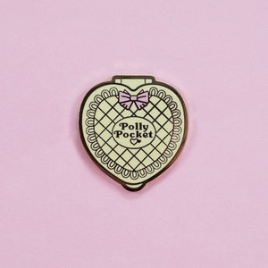 Puede incluir: Pin compacto de Polly Pocket en forma de coraz&oacute;n con borde dorado, interior verde claro y lazo rosa. El pin presenta las palabras "Polly Pocket" en un dise&ntilde;o circular. El fondo es de color rosa claro liso.