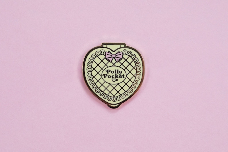 Puede incluir: Pin compacto de Polly Pocket en forma de coraz&oacute;n con borde dorado, interior verde claro y lazo rosa. El pin presenta las palabras "Polly Pocket" en un dise&ntilde;o circular. El fondo es de color rosa claro liso.