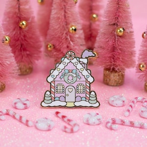 May include: A pink and white gingerbread house enamel pin with candy cane details. The pin is surrounded by pink miniature Christmas trees and candy canes on a pink glitter background.