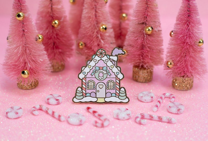 May include: A pink and white gingerbread house enamel pin with candy cane details. The pin is surrounded by pink miniature Christmas trees and candy canes on a pink glitter background.