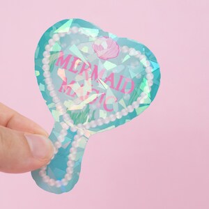 Puede incluir: Un espejo de mano de corazón turquesa con un borde de perlas. El espejo tiene un acabado holográfico y el texto "Mermaid Magic" en letras rosas.