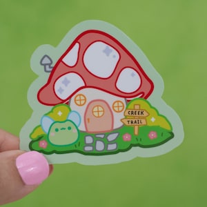 May include: A sticker featuring a cartoon mushroom house with a pink door, a green frog with wings, and a sign that reads "Creek Trail".