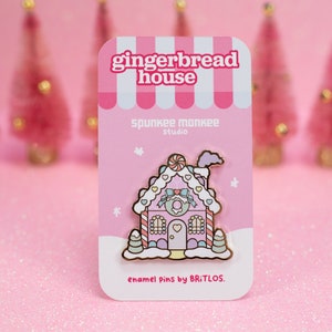 May include: Pink gingerbread house enamel pin with white frosting, candy canes, and a heart-shaped door. The pin is on a pink card with the text "gingerbread house", "spunkee monkee studio", and "enamel pins by BRITLOS."