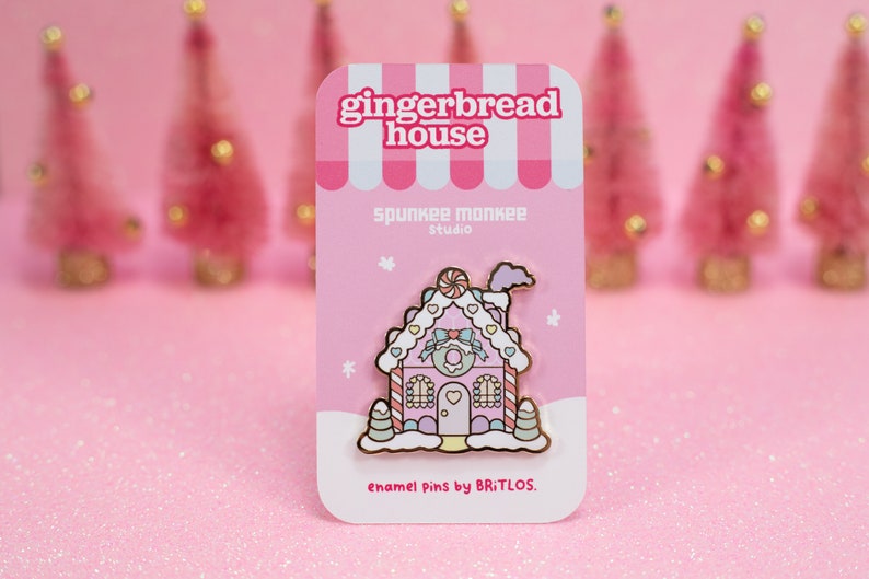 May include: Pink gingerbread house enamel pin with white frosting, candy canes, and a heart-shaped door. The pin is on a pink card with the text "gingerbread house", "spunkee monkee studio", and "enamel pins by BRITLOS."