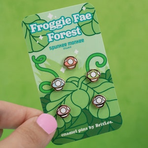 K&ouml;nnte beinhalten: Satz mit f&uuml;nf Emaille-Pins mit verschiedenen farbigen Pilzen mit wei&szlig;en Punkten. Die Pins befinden sich auf einer Karte mit dem Text "Froggy Fae Forest" und "enamel pins by BritLos."