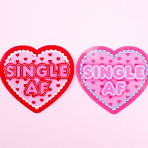 Op de afbeelding: Twee roze en rode hartvormige decoraties met de tekst "SINGLE AF" in witte letters. De harten hebben een witte gegolfde rand en een patroon van kleine roze hartjes.