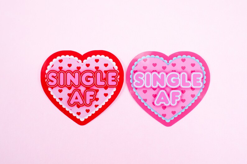 Op de afbeelding: Twee roze en rode hartvormige decoraties met de tekst "SINGLE AF" in witte letters. De harten hebben een witte gegolfde rand en een patroon van kleine roze hartjes.