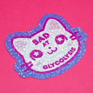 May include: A glittery sticker in the shape of a cat's head, with a blue outline and a white center. The sticker has the words "BAD AT GLYCOLYSIS" in purple. The background is a solid pink color.