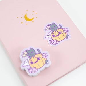 May include: Two stickers featuring a cartoon pumpkin with a cat sitting on top. The pumpkin is yellow and the cat is gray. The stickers are on a pink background with a gold moon and stars.