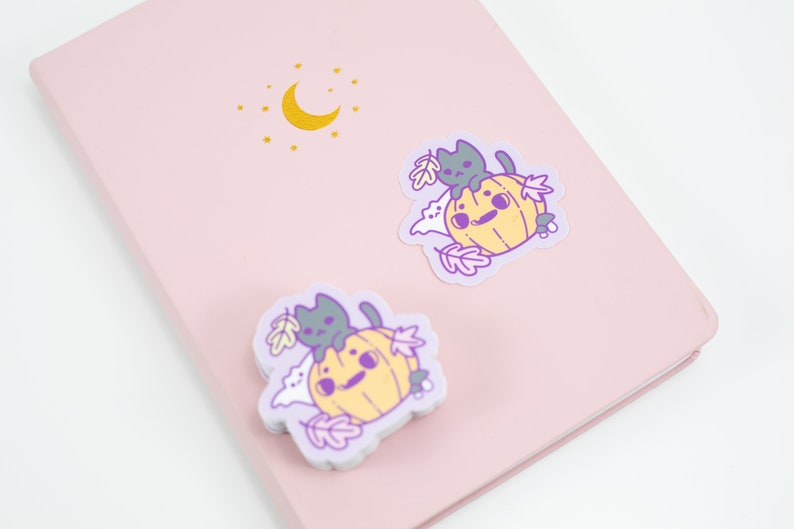 May include: Two stickers featuring a cartoon pumpkin with a cat sitting on top. The pumpkin is yellow and the cat is gray. The stickers are on a pink background with a gold moon and stars.