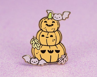 Op de afbeelding: Een gouden emaille pin met een stapel van drie glimlachende pompoenen met een vleermuis erop. De pompoenen zijn oranje met zwarte ogen en een zwarte mond. De vleermuis is paars met witte ogen en een zwarte neus.