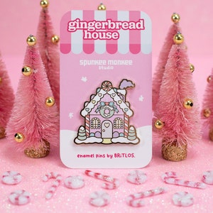 May include: A pink gingerbread house enamel pin with white frosting, candy canes, and a pink bow. The pin is on a pink card with the text "gingerbread house" and "Spunkee Monkee Studio" in white. The card also says "enamel pins by Britlos."