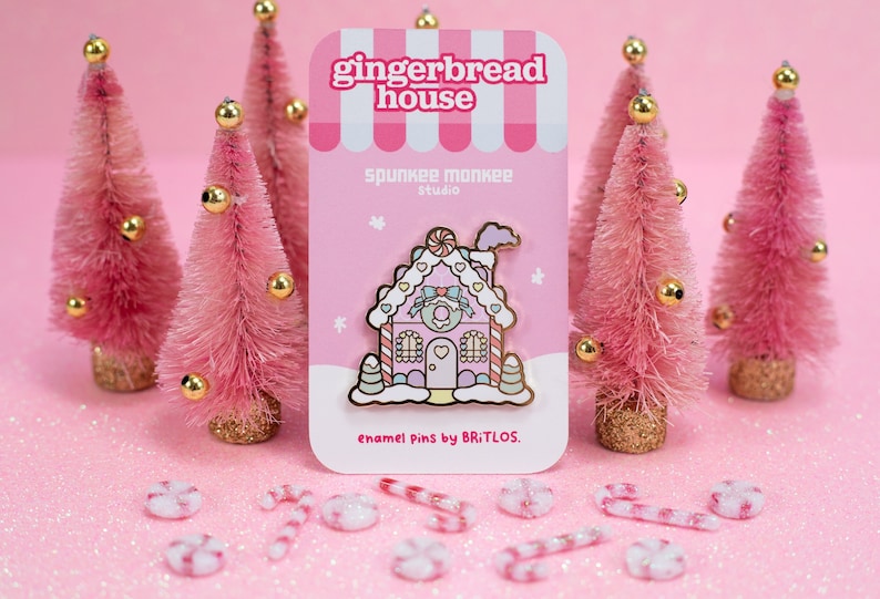 May include: A pink gingerbread house enamel pin with white frosting, candy canes, and a pink bow. The pin is on a pink card with the text "gingerbread house" and "Spunkee Monkee Studio" in white. The card also says "enamel pins by Britlos."