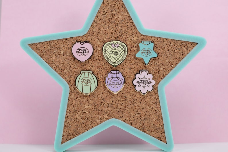 Puede incluir: Seis pines esmaltados de Polly Pocket en tonos pastel, con formas de coraz&oacute;n, estrella y flor. Cada pin presenta el logotipo de Polly Pocket. Se exhiben sobre un corcho dentro de un marco en forma de estrella turquesa.