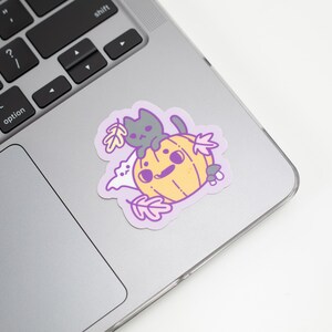 May include: A sticker featuring a cartoon cat sitting on a pumpkin with a ghost and leaves. The sticker is on a silver laptop.