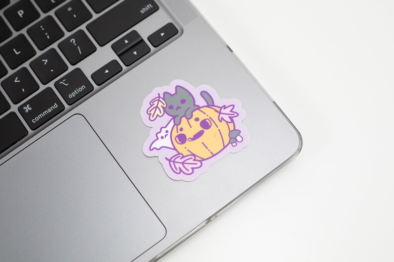 May include: A sticker featuring a cartoon cat sitting on a pumpkin with a ghost and leaves. The sticker is on a silver laptop.