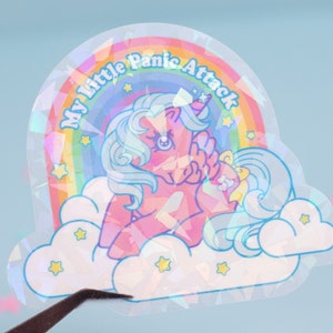 May include: A holographic sticker featuring a pink and blue cartoon unicorn with a rainbow and stars. The sticker says "My little Panic Attack".