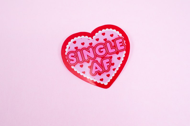 Op de afbeelding: Een rode hartvormige kaart met een roze achtergrond en witte gegolfde randen. De kaart zegt "SINGLE AF" in roze letters.