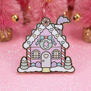 May include: A pink and white gingerbread house enamel pin with candy cane details, a candy cane on the roof, and a heart-shaped door. The pin is surrounded by pink glitter.