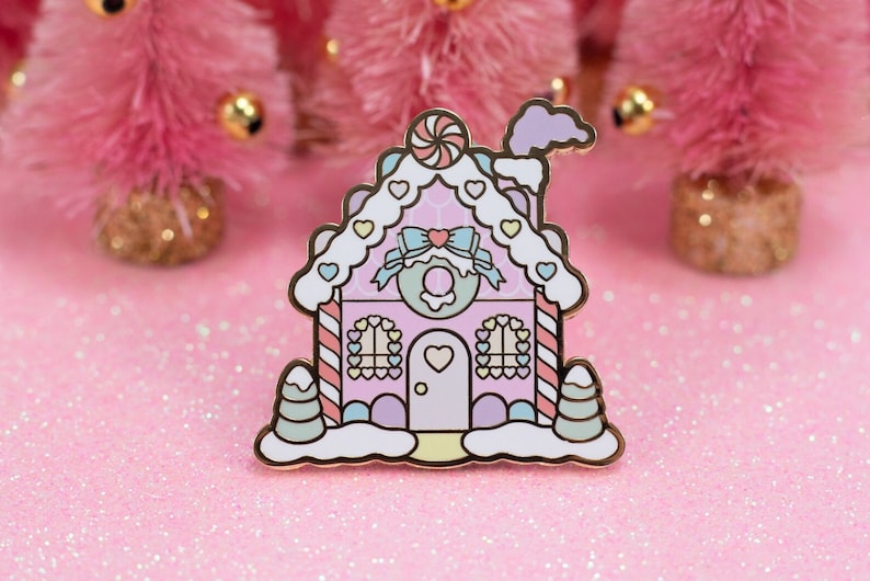 May include: A pink and white gingerbread house enamel pin with candy cane details, a candy cane on the roof, and a heart-shaped door. The pin is surrounded by pink glitter.
