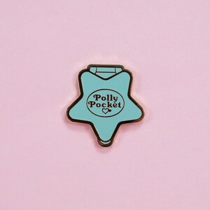 Puede incluir: Un pin de Polly Pocket en forma de estrella turquesa con borde dorado, con el texto "Polly Pocket" y un coraz&oacute;n. El pin est&aacute; sobre un fondo rosa suave, creando una est&eacute;tica l&uacute;dica y nost&aacute;lgica.
