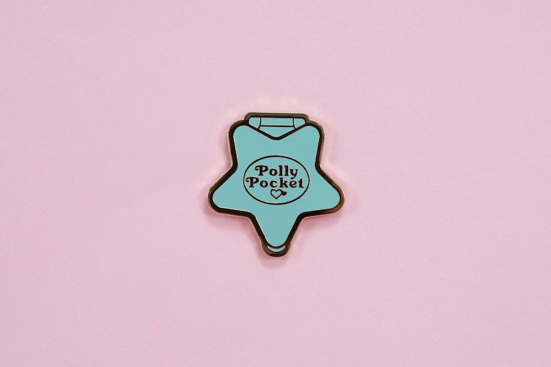 Puede incluir: Un pin de Polly Pocket en forma de estrella turquesa con borde dorado, con el texto "Polly Pocket" y un coraz&oacute;n. El pin est&aacute; sobre un fondo rosa suave, creando una est&eacute;tica l&uacute;dica y nost&aacute;lgica.