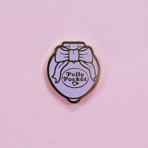 Puede incluir: Un pin de esmalte de Polly Pocket de color lavanda con ribetes dorados. El pin presenta un lazo en la parte superior y las palabras "Polly Pocket" en un dise&ntilde;o en forma de coraz&oacute;n. El pin est&aacute; sobre un fondo rosa.