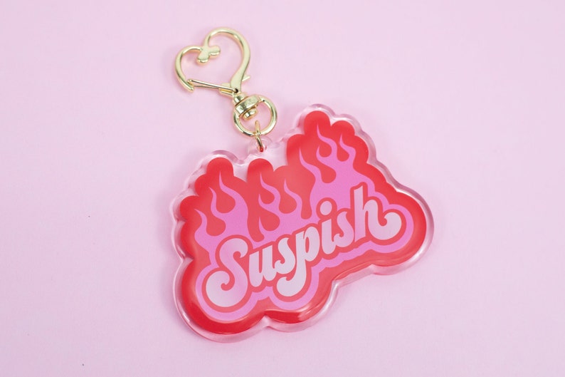 Pode incluir: Um chaveiro de acr&iacute;lico vermelho e rosa com um design de chama e a palavra "Suspish" em letras brancas.