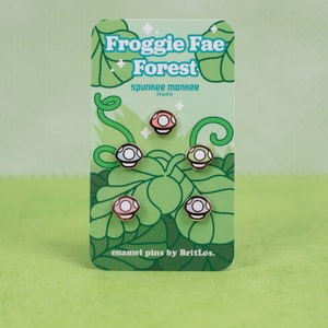K&ouml;nnte beinhalten: Ein Set mit f&uuml;nf Emaille-Pins, die verschiedene farbige Pilze mit wei&szlig;en Punkten zeigen. Die Pins sind auf einer Karte verpackt, auf der der Text "Froggie Fae Forest" und "spunkie monkee studio" in einem gr&uuml;n-wei&szlig;en Farbschema steht. Die Karte enth&auml;lt auch den Text "enamel pins by BritLos."