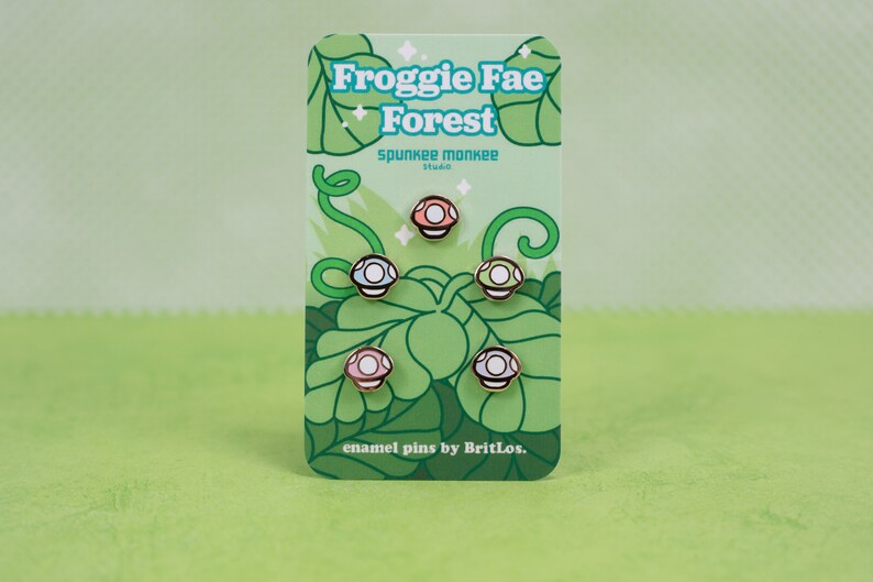 K&ouml;nnte beinhalten: Ein Set mit f&uuml;nf Emaille-Pins, die verschiedene farbige Pilze mit wei&szlig;en Punkten zeigen. Die Pins sind auf einer Karte verpackt, auf der der Text "Froggie Fae Forest" und "spunkie monkee studio" in einem gr&uuml;n-wei&szlig;en Farbschema steht. Die Karte enth&auml;lt auch den Text "enamel pins by BritLos."