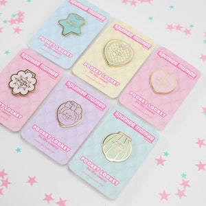 Puede incluir: Una colecci&oacute;n de pines Pocket Locket de Spunkee Monkee en dorado, con dise&ntilde;os como una estrella, un coraz&oacute;n, una flor, un lazo y una concha. Cada pin se presenta en una tarjeta de color pastel con el texto "Pocket Locket".