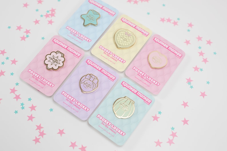 Puede incluir: Una colecci&oacute;n de pines Pocket Locket de Spunkee Monkee en dorado, con dise&ntilde;os como una estrella, un coraz&oacute;n, una flor, un lazo y una concha. Cada pin se presenta en una tarjeta de color pastel con el texto "Pocket Locket".