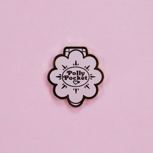 Puede incluir: Un pin de esmalte rosa claro con forma de flor y contorno negro. El pin presenta las palabras "Polly Pocket" en un dise&ntilde;o circular con un coraz&oacute;n en el centro. El fondo es de color rosa claro liso.