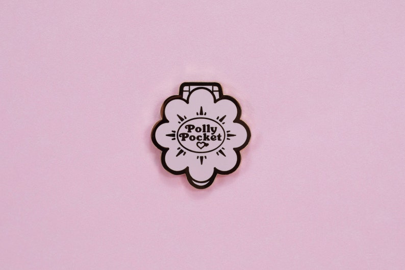 Puede incluir: Un pin de esmalte rosa claro con forma de flor y contorno negro. El pin presenta las palabras "Polly Pocket" en un dise&ntilde;o circular con un coraz&oacute;n en el centro. El fondo es de color rosa claro liso.