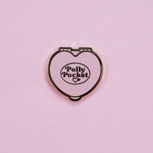 Puede incluir: Un compacto Polly Pocket en forma de coraz&oacute;n rosa claro con borde dorado. El compacto presenta las palabras "Polly Pocket" en una fuente retro, con un peque&ntilde;o s&iacute;mbolo de coraz&oacute;n debajo. El fondo es de color rosa claro liso.