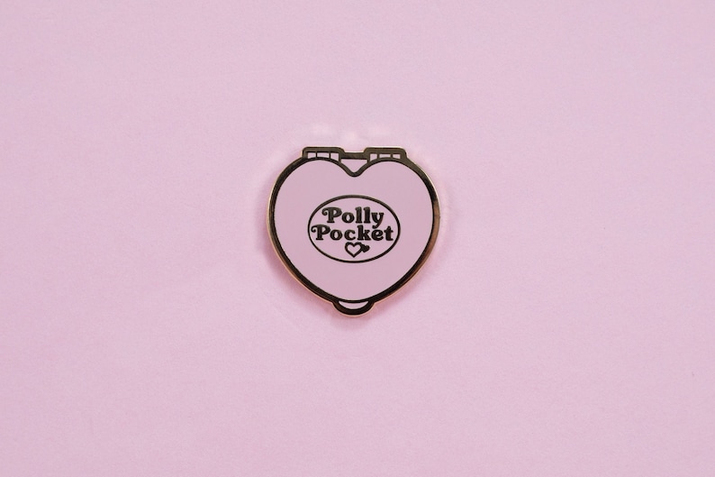 Puede incluir: Un compacto Polly Pocket en forma de coraz&oacute;n rosa claro con borde dorado. El compacto presenta las palabras "Polly Pocket" en una fuente retro, con un peque&ntilde;o s&iacute;mbolo de coraz&oacute;n debajo. El fondo es de color rosa claro liso.